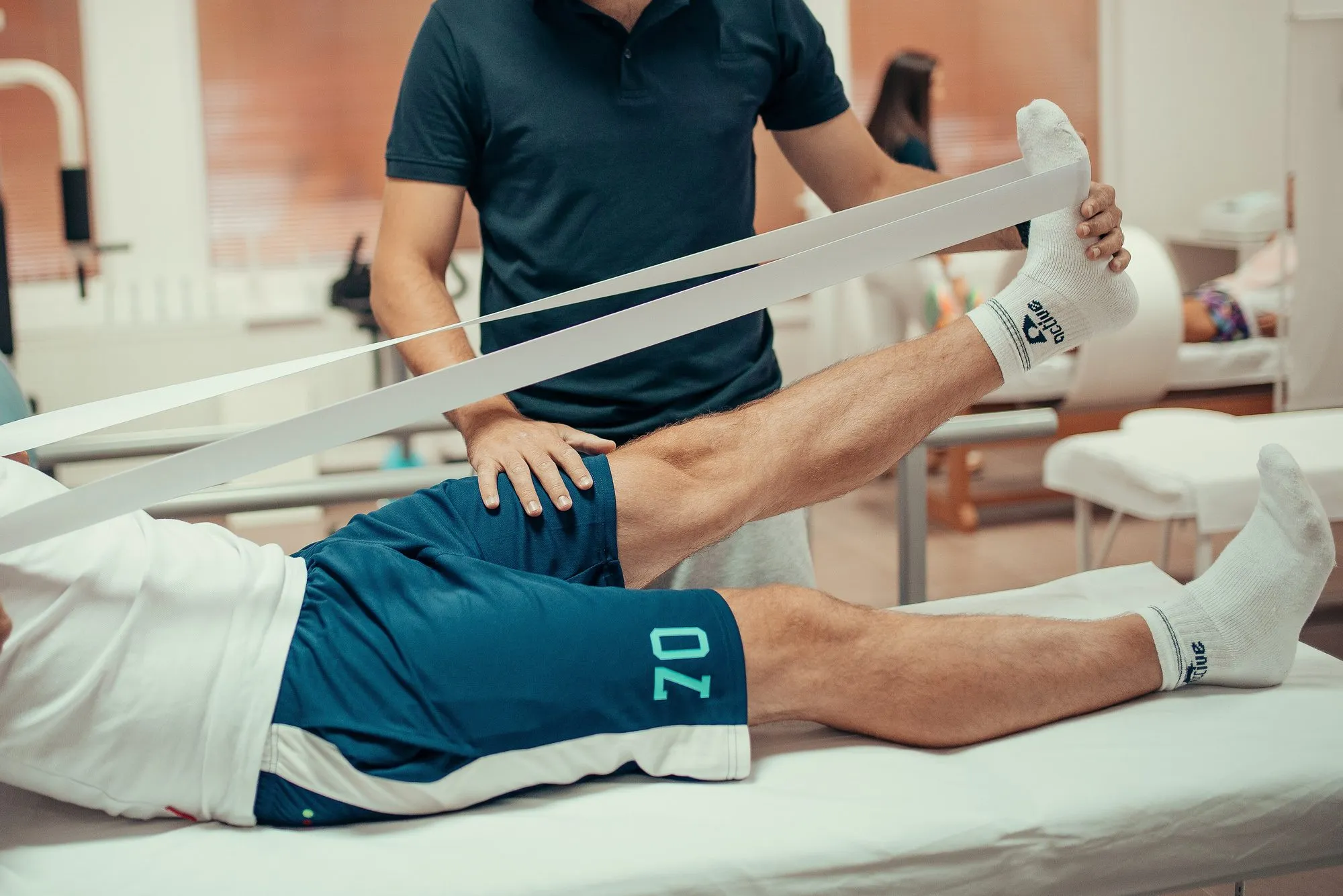 Ein Patient hebt auf der Liege das gestreckte Bein gegen ein Gymnastikband, der Therapeut stützt den Oberschenkel