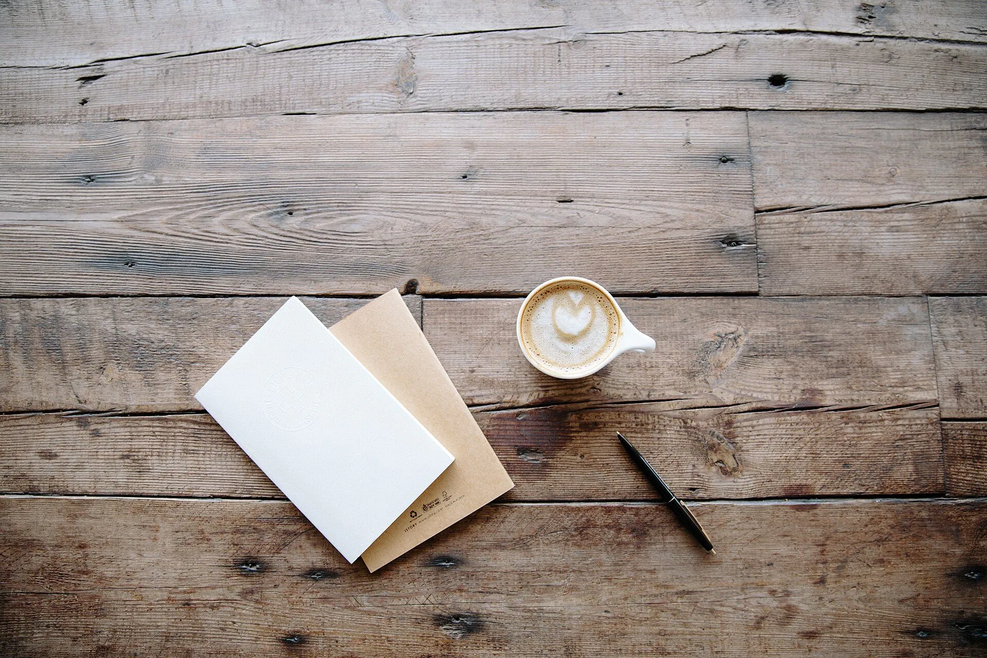 Zwei Notizbücher, ein Stift und ein Cappuccino auf einem alten Holztisch