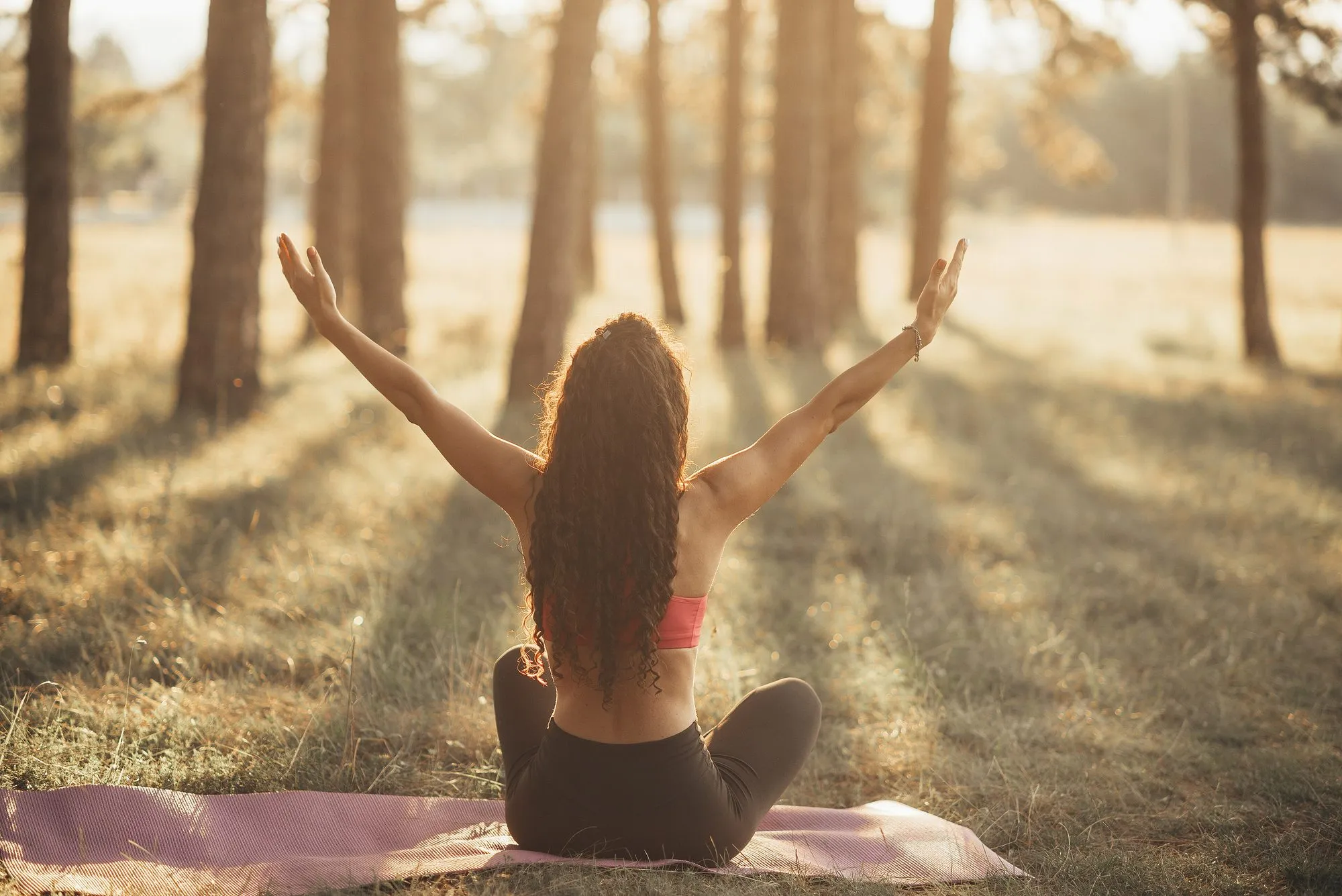 Frau im Schneidersitz auf einer Yogamatte im Park, die Arme in die Abendsonne gestreckt